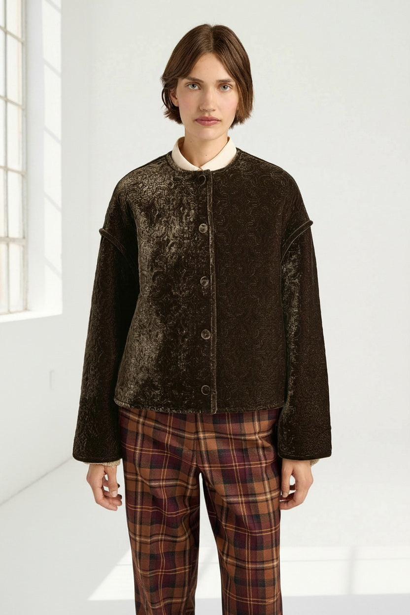 A person stands in a bright room wearing the MOMONI - CRANBERRY QUILTED COAT IN BROWN over a collared shirt and plaid pants, looking directly at the camera.