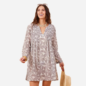 A woman in the ROCKFLOWERPAPER PALM LEAVES GREY PINTUCK COVERUP smiles and looks to the side, holding a straw hat, with layered necklaces and sunglasses on her head.