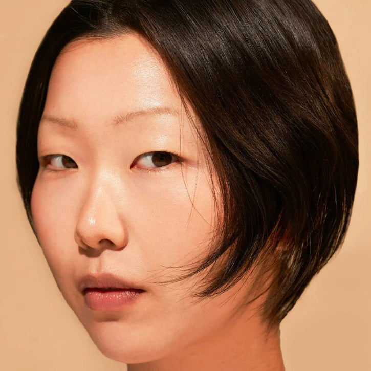 A close-up of a person with natural curly hair, adorned in gold hoop earrings and a white top. They wear a neutral facial expression while looking directly at the camera. The softly lit, plain background enhances their glowing skin as if touched by JASON WU - TINTED MOISTURIZER MEETS CC CREAM from JASON WU BEAUTY.