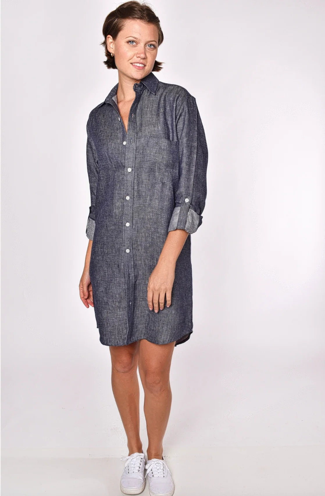 A person stands against a plain light background, smiling, in the EMILY PHILLIPS - THE LINEN DENIM MINI SHIRTDRESS from EMILY PHILLIPS—a long-sleeve, button-down dress made from sustainable linen with a dark chambray look—styled with rolled-up sleeves and white sneakers.
