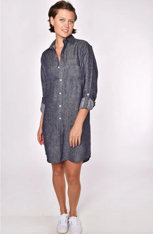 A person stands against a plain light background, smiling, in the EMILY PHILLIPS - THE LINEN DENIM MINI SHIRTDRESS from EMILY PHILLIPS—a long-sleeve, button-down dress made from sustainable linen with a dark chambray look—styled with rolled-up sleeves and white sneakers.