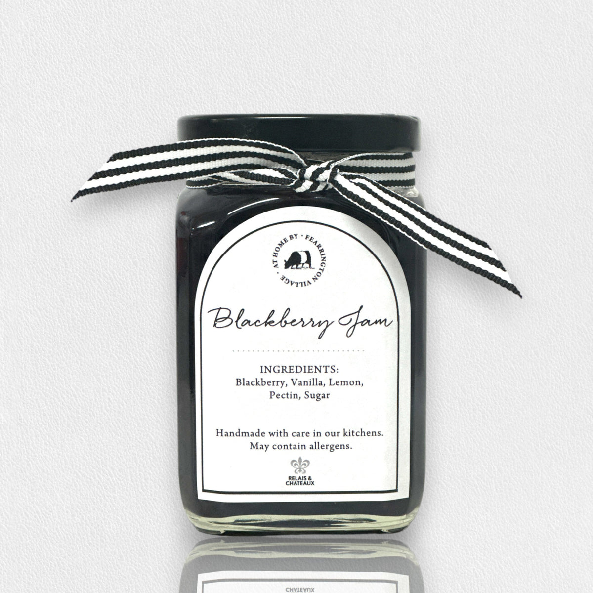 The FEARRINGTON SIGNATURE COLLECTION - FEARRINGTON HOUSEMADE BLACKBERRY JAM features a stylish black and white striped ribbon tied around the lid. The label reveals ingredients including blackberry, vanilla, lemon, pectin, and sugar. This handcrafted jam is made with meticulous care and is an ideal hostess gift. Please be aware that it may contain allergens.