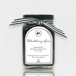 The FEARRINGTON SIGNATURE COLLECTION - FEARRINGTON HOUSEMADE BLACKBERRY JAM features a stylish black and white striped ribbon tied around the lid. The label reveals ingredients including blackberry, vanilla, lemon, pectin, and sugar. This handcrafted jam is made with meticulous care and is an ideal hostess gift. Please be aware that it may contain allergens.