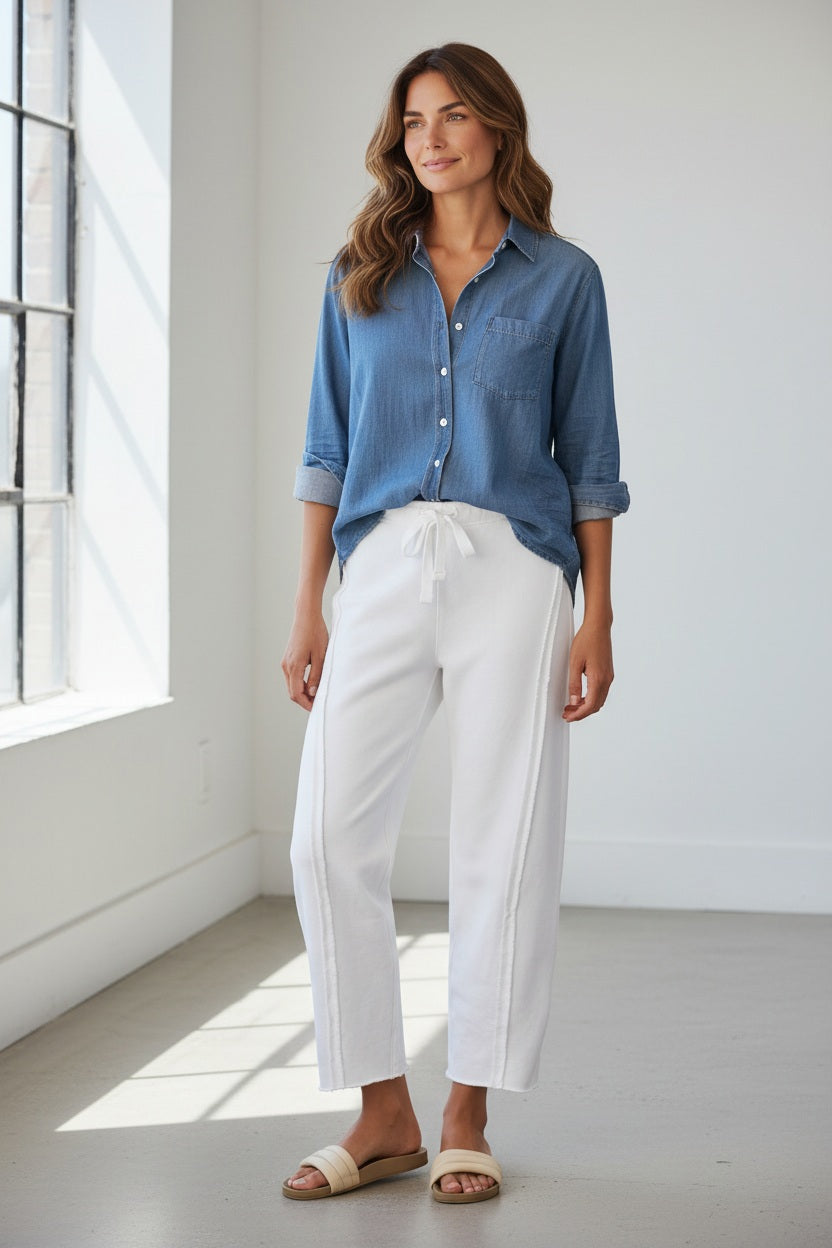 A person wearing the FRANK &amp; EILEEN Bibi Barrel Leg Sweatpant in Triple Fleece White stands on a patio next to potted plants with pink flowers, paired with a blue long-sleeve shirt and beige sandals.