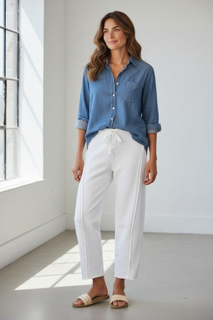 A person wearing the FRANK & EILEEN Bibi Barrel Leg Sweatpant in Triple Fleece White stands on a patio next to potted plants with pink flowers, paired with a blue long-sleeve shirt and beige sandals.