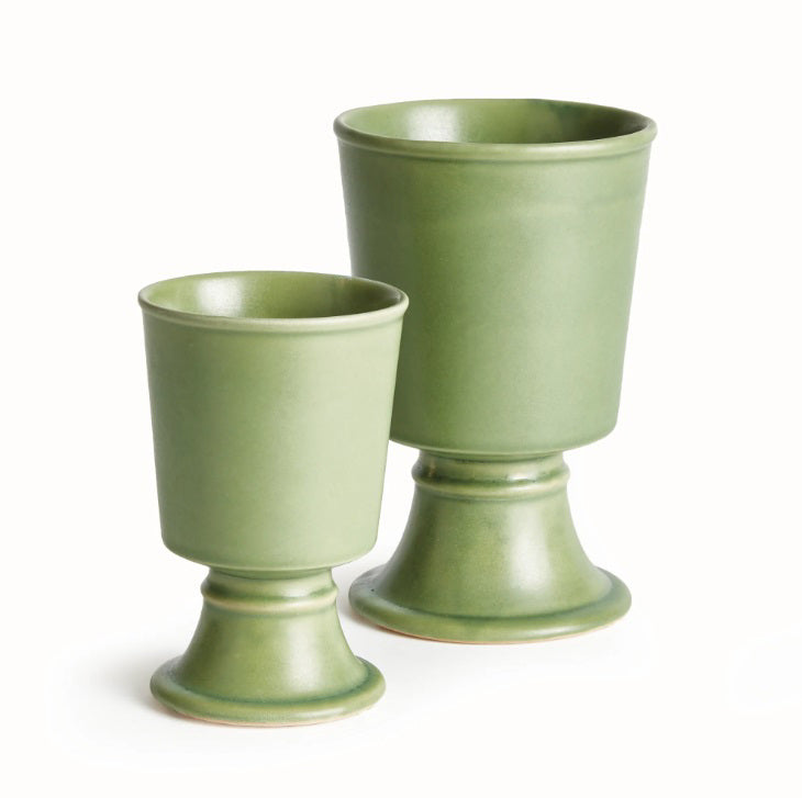 Two NAPA HOME AND GARDEN - KELCY FOOTED CACHEPOTS, finished with a green reactive glaze, rest on a light woven runner. One displays a leafy plant while the other is empty; a wooden chair and white slatted fence are seen in the background.