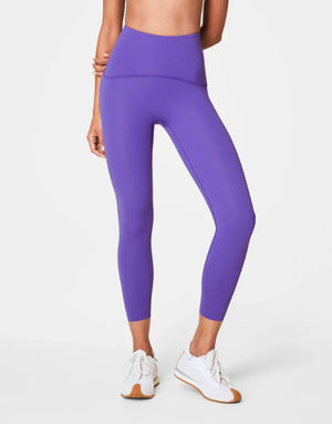 A person wears SPANX - BOOTY BOOST 7/8 LEGGINGS in high-waisted royal blue performance fabric with black athletic shoes, standing hand on hip against a plain white background. Only their lower body is visible; upper body and face are not shown.