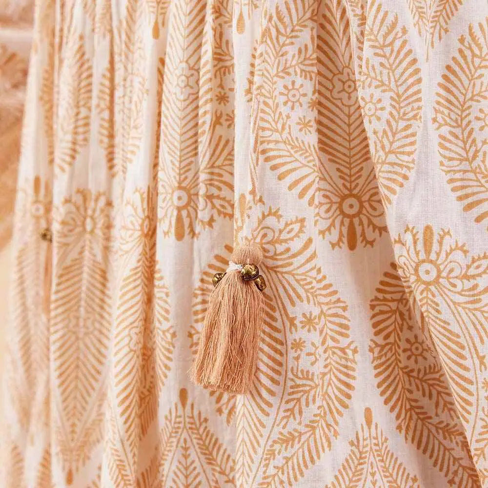 A woman smiles and looks down while wearing the ROCKFLOWERPAPER - ZAHARRA DRESS, a lightweight cream long-sleeve coverup with a subtle pattern and tassels, standing against a beige background.