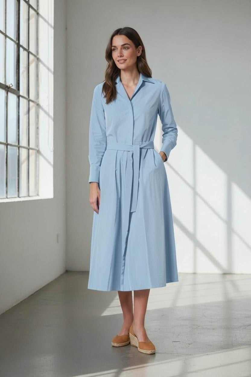 A woman stands indoors in sunlight streaming through large windows, wearing the VICARIO CINQUE Anna Taffeta Shirtdress in Light Blue—a long-sleeve memory taffeta dress with a belted waist and side pockets—paired with tan espadrille shoes.