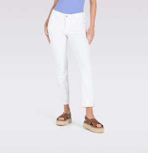 With long brown hair, a person stands against a plain backdrop wearing MAC's tan cropped jacket and MAC JEANS - DREAM SUMMER light blue slim jeans made from a comfortable modal blend. White heels complete the look as they gaze ahead with relaxed arms by their sides.