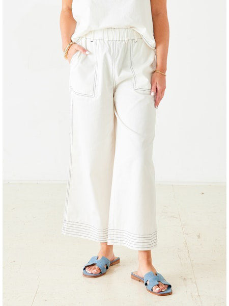 A person stands indoors wearing MERSEA - SAMMIE WIDE LEG PANT, relaxed-fit high-waisted white cotton pants with visible stitching. They pair them with a white top, gold bracelets, and blue slide sandals; only their lower body and hands are visible.