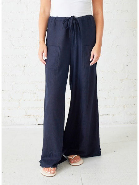 A person wearing a white sleeveless top, MERSEA - PROVENCE PALAZZO PANT in navy blue with front pockets and a drawstring waist, and white flip-flops stands against a white brick wall.