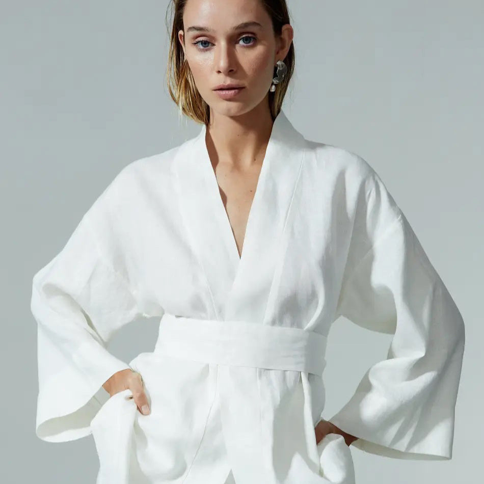 A woman with light hair wears a white, kimono-style robe with wide sleeves and a matching belt, standing with her hands in her pockets against a plain gray background.