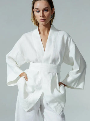 A woman stands against a plain background, wearing a white, kimono-style wrap top with wide sleeves and a matching belt, her hands in the garment’s large pockets.