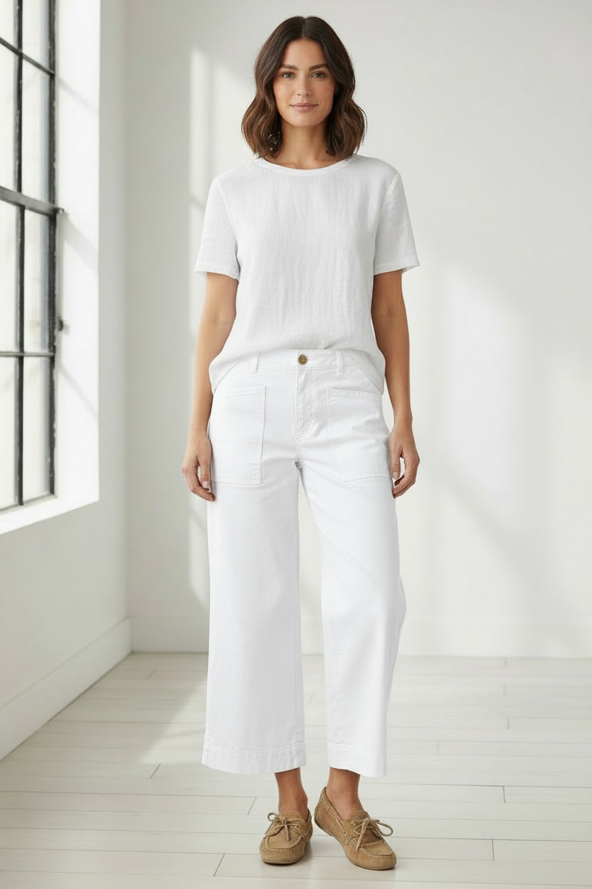 A person wearing the FRANK &amp; EILEEN Limerick Dropped Wide Leg Carpenter Pant in Italian Performance Linen White, paired with a blue and white striped top and flip-flops, stands on sandy ground between two white walls with the ocean visible in the background. The cropped length of these FRANK &amp; EILEEN pants enhances the breezy, relaxed feel of the seaside scene.