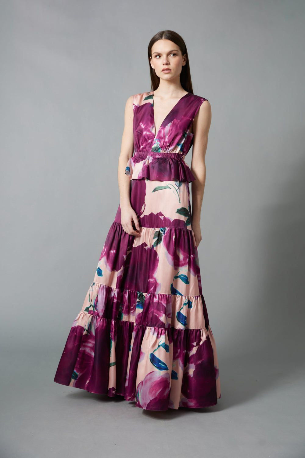 A woman with straight brown hair and a neutral expression wears the HOPE FOR FLOWERS Tiered Maxi Dress, a sleeveless, floor-length style featuring bold purple and pink floral print, standing against a gray background.