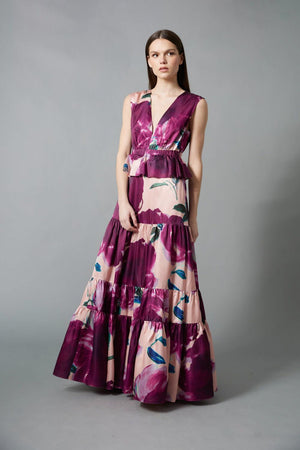A woman with straight brown hair and a neutral expression wears the HOPE FOR FLOWERS Tiered Maxi Dress, a sleeveless, floor-length style featuring bold purple and pink floral print, standing against a gray background.