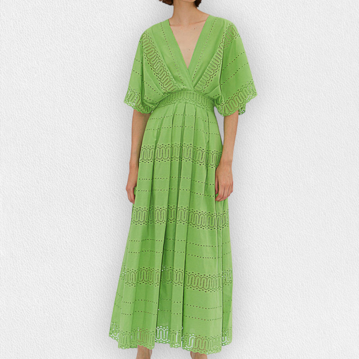 A person wearing the MILVA SANGALLO DRESS, a light green, full-length dress with a deep V-neckline and intricate eyelet lace details, perfect for a garden party. The dress features short, flared sleeves and a flowy skirt with horizontal eyelet patterns. The person is standing against a plain, light-textured background.