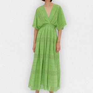 A person wearing the MILVA SANGALLO DRESS, a light green, full-length dress with a deep V-neckline and intricate eyelet lace details, perfect for a garden party. The dress features short, flared sleeves and a flowy skirt with horizontal eyelet patterns. The person is standing against a plain, light-textured background.