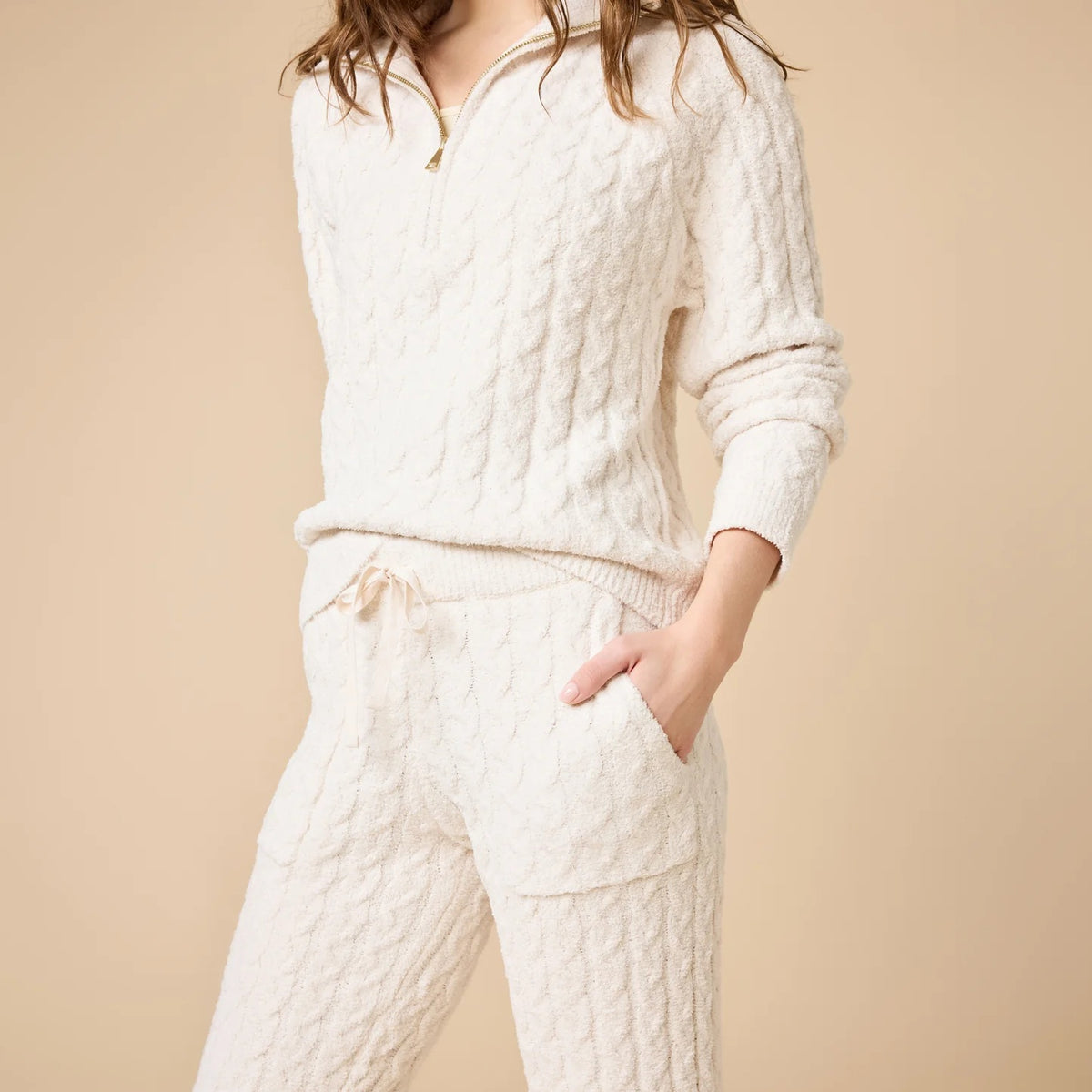 A woman wearing a marshmallow-soft, cream-colored SOFTIES - CABLE KNIT MARSHMALLOW ½ ZIP WITH TAPERED PANT SET, featuring a half-zip pullover and matching pants with a drawstring waist.