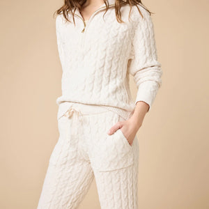 A woman wearing a marshmallow-soft, cream-colored SOFTIES - CABLE KNIT MARSHMALLOW ½ ZIP WITH TAPERED PANT SET, featuring a half-zip pullover and matching pants with a drawstring waist.