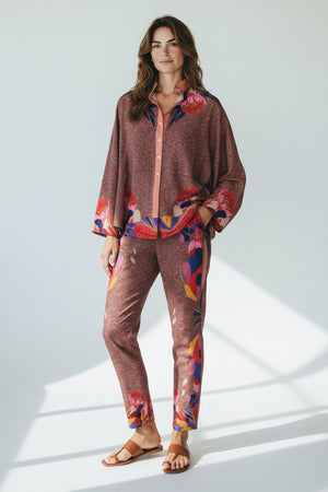 A person stands indoors wearing FRANCO FERRARI - SILK PRINT TROUSER, featuring loose-fitting silk with colorful abstract and subtle floral designs in brown, pink, and blue. They pair it with gold shoes, posed before sheer draped fabric and wooden objects.