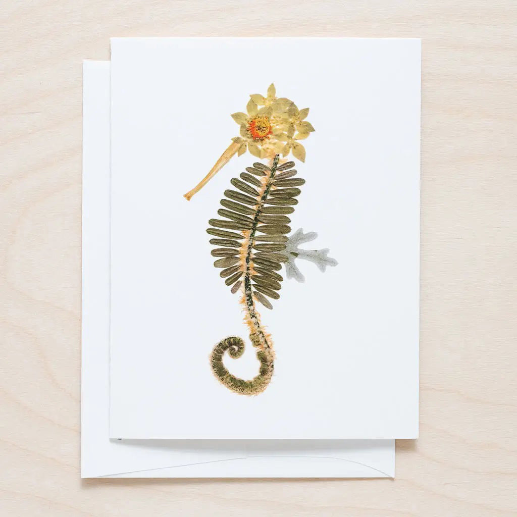 The PETAL PEOPLE PRESS - SEAHORSE CARD features a seahorse design made from pressed flowers and leaves, with green leaves for the body and yellow-brown flowers for the head and tail, set against a light wooden background.