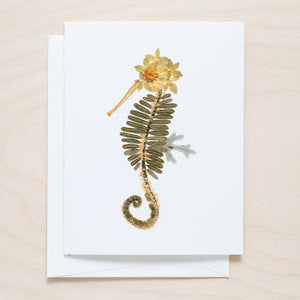 The PETAL PEOPLE PRESS - SEAHORSE CARD features a seahorse design made from pressed flowers and leaves, with green leaves for the body and yellow-brown flowers for the head and tail, set against a light wooden background.