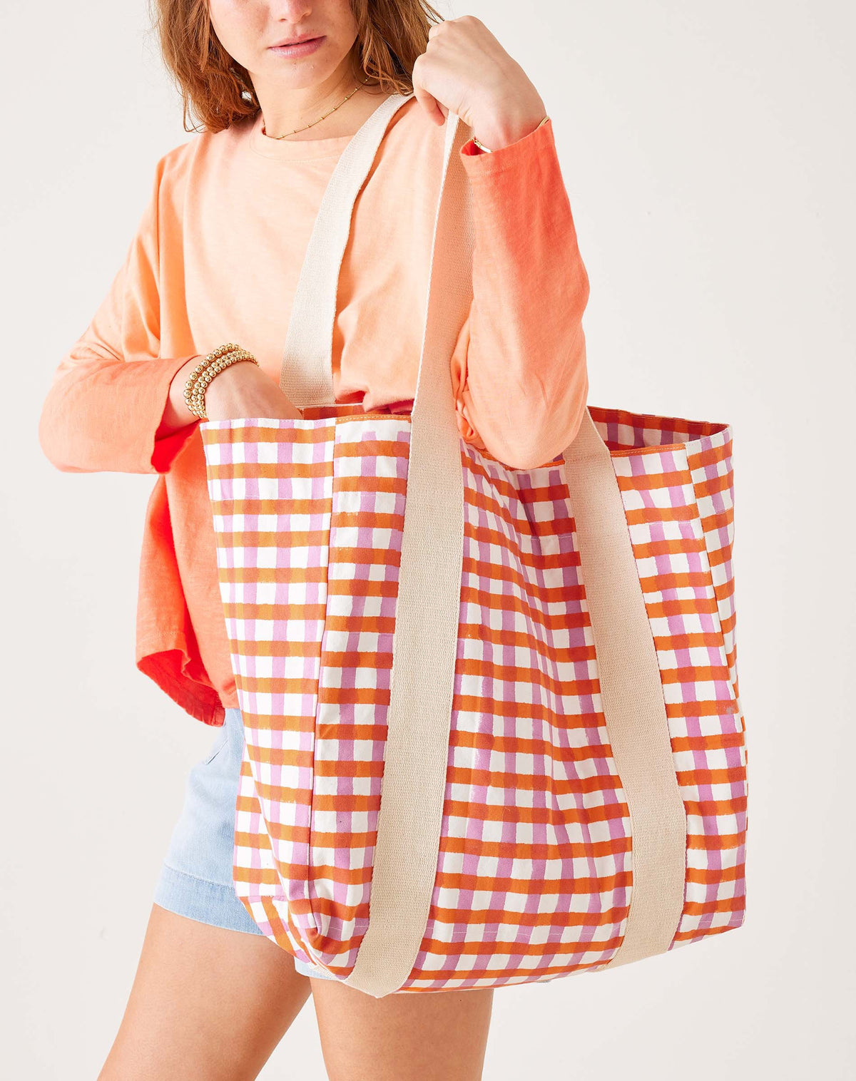 A woman in an orange long-sleeve shirt and denim shorts carries the MERSEA LEO TOTE BAG, a high-quality cotton tote from MERSEA with wide cream straps and an open top, featuring colorful orange, pink, and white checkered squares.