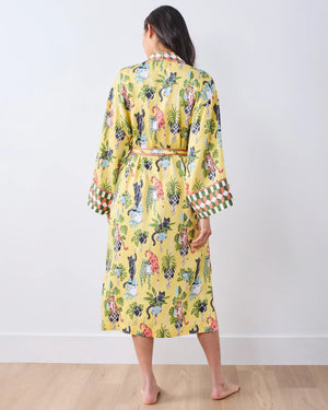 A woman stands barefoot against a white wall, wearing the PRINTFRESH - Macrame Mischief Eco Satin Robe in Sunshine—a long yellow robe featuring colorful animal and botanical designs, geometric trim, and a matching belt—perfect for any cat lover.