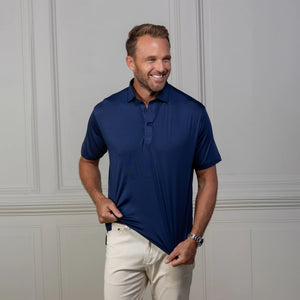 A man in the COLLARS AND CO FEATHERWEIGHT FLEX POLO IN NAVY and white pants stands by a light-paneled wall, smiling and holding the side of his performance golf shirt.