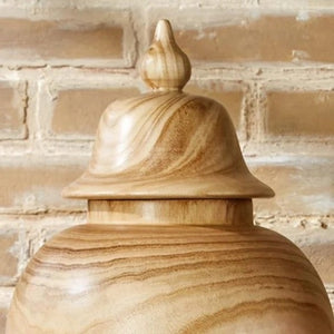 The PAULOWNIA WOOD COVERED TEMPLE JAR - LARGE by TWO'S COMPANY rests elegantly on a wooden surface before a light brick wall, its tall silhouette and swirling grain patterns enhanced by a fitted lid. Green plant leaves peeking in from the right complete this exotic home accessory, bringing warmth and character to any space.