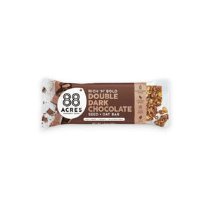 An 88 ACRES - DOUBLE DARK CHOCOLATE SEED + OAT BAR rests on a light wooden surface. The packaging is brown and white, showcasing images of dark chocolate chunks and oats. The label proudly highlights that it is gluten-free, vegan, and nut-free.