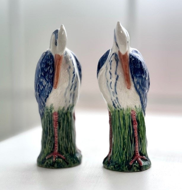 The QUAIL DESIGNS QUAIL CERAMICS Heron Salt and Pepper Shakers feature two hand-painted blue and white herons on green grass, facing each other. Dishwasher safe for easy cleaning.
