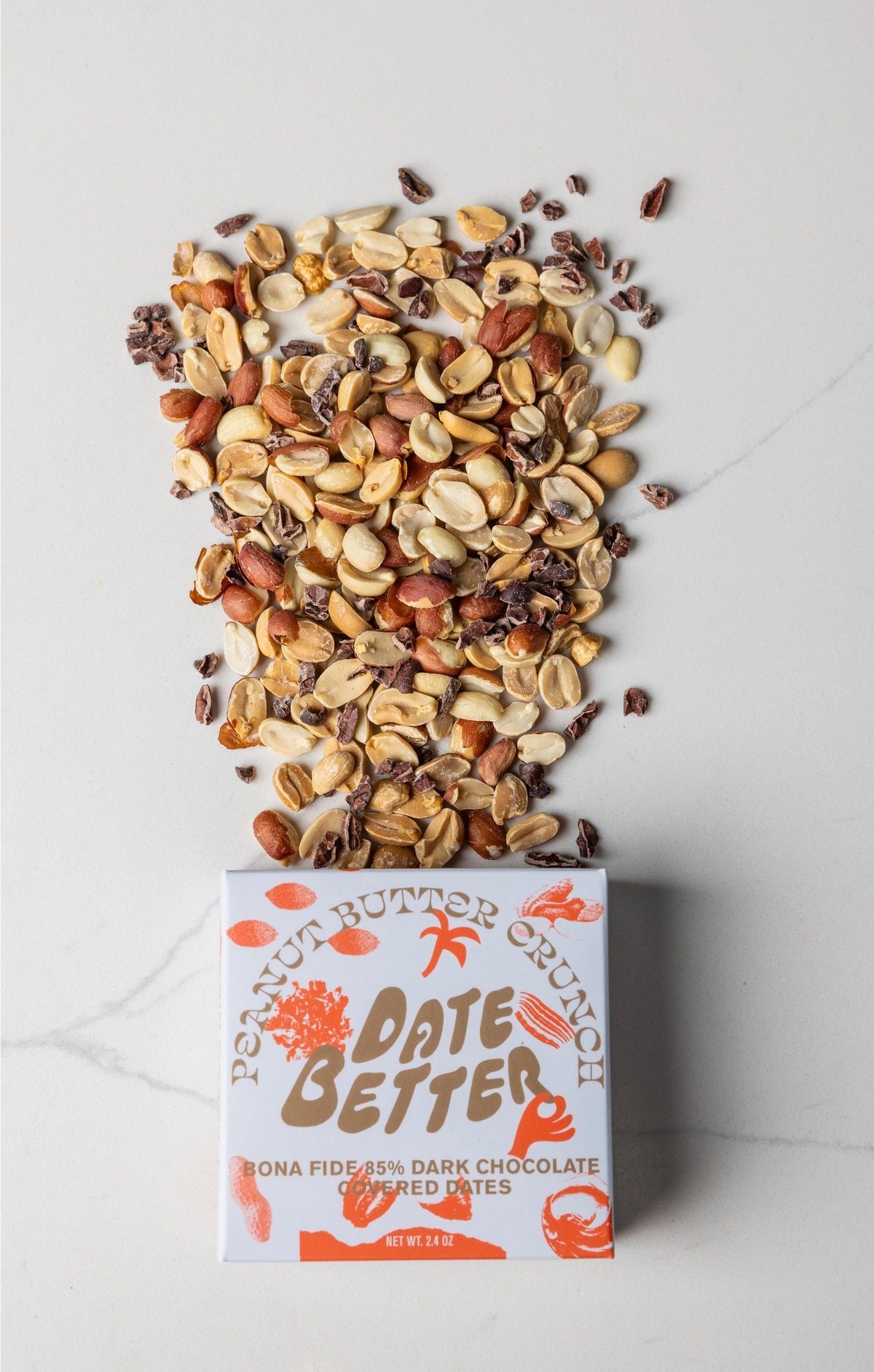 A white "DATE BETTER - PEANUT BUTTER CRUNCH COVERED DATES" package from DATE BETTER SNACKS rests on marble, cacao nibs and nuts spilling out. Orange text and illustrations adorn the packaging, adding charm to the scene.