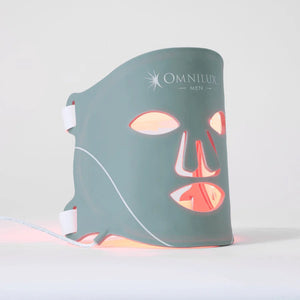 A man intently examines the OMNILUX - MENS MASK, a glowing red LED mask designed for skin rejuvenation with advanced red light therapy, featuring eye, nose, and mouth cutouts.