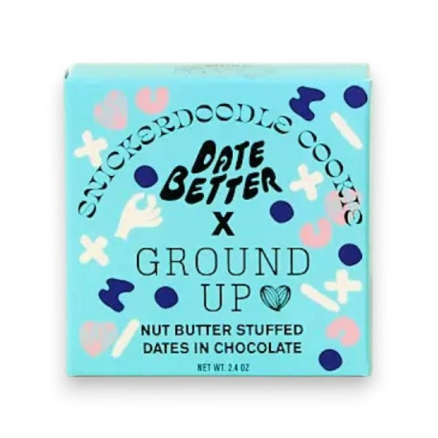 A box of DATE BETTER SNACKS’ DATE BETTER - SNICKERDOODLE COOKIE features nut butter stuffed dates enrobed in dark chocolate, artfully arranged with nuts, a cinnamon stick, and a bitten cookie to showcase the ultimate Snickerdoodle Cookie Crunch—all presented on a pastel backdrop.