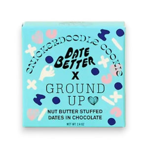 A box of DATE BETTER SNACKS’ DATE BETTER - SNICKERDOODLE COOKIE features nut butter stuffed dates enrobed in dark chocolate, artfully arranged with nuts, a cinnamon stick, and a bitten cookie to showcase the ultimate Snickerdoodle Cookie Crunch—all presented on a pastel backdrop.