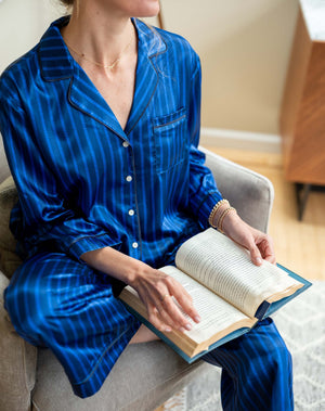 The MERSEA - Carousel Stripe Satin Sailor Pajama Set in Blue by MERSEA, featuring elegant blue and black stripes with a button-up top and matching pants, embodies luxury sleepwear and is neatly folded on a light blue background.