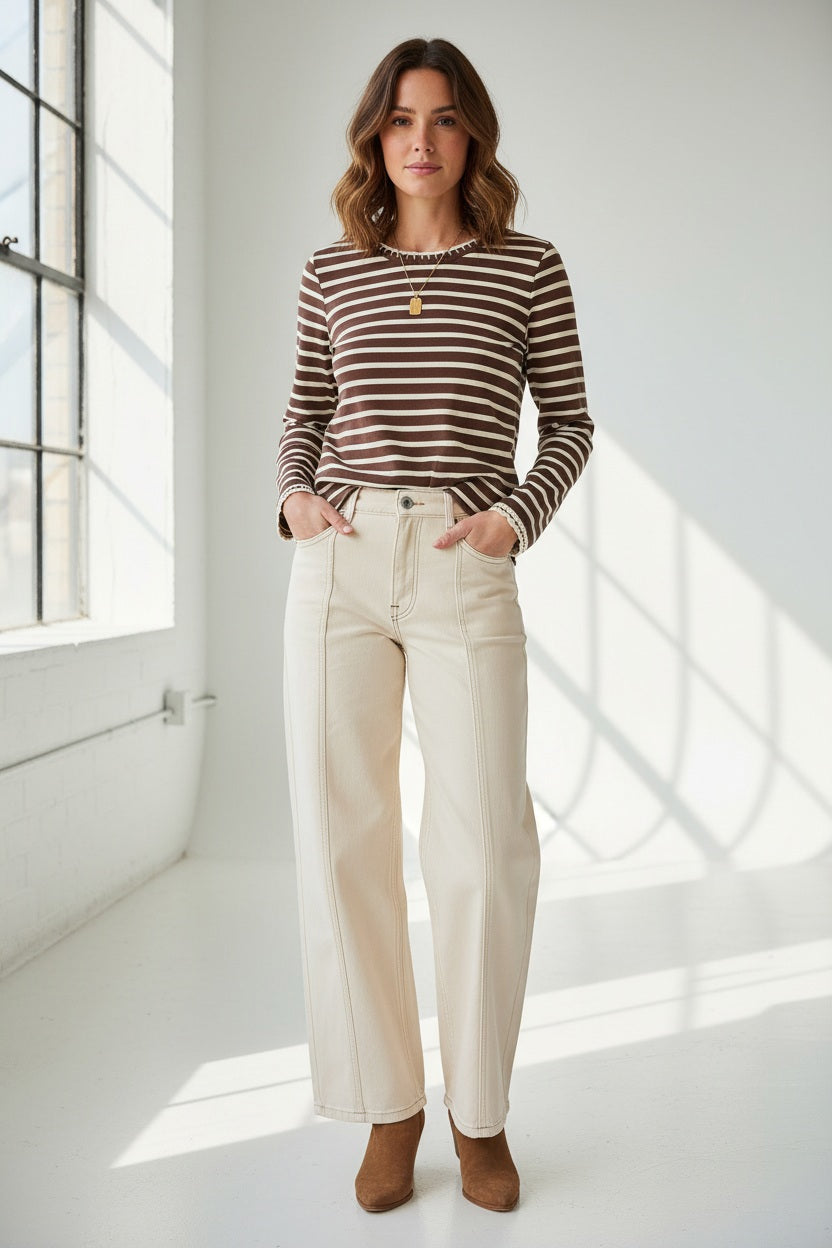 A woman stands in a bright room wearing the MERSEA Mariner Tee in Espresso Creme Stripe, paired with cream wide-leg pants and brown boots, her hands tucked into her pockets.