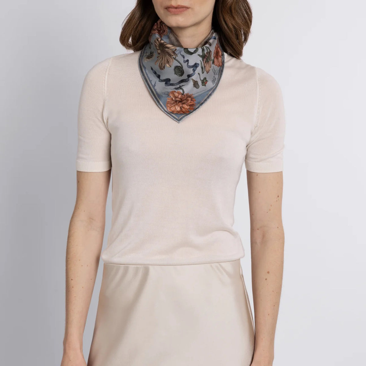 A woman holds the SABINA SAVAGE "Ring a Ring o' Roses" silk twill scarf in Glacier/Ballet, featuring a blue-eyed cat with a ribbon amidst Victorian botanicals and a historic building backdrop, with "Ring a Ring o' Roses, A Pocket Full of Posies" elegantly displayed.