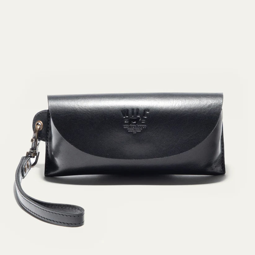 The WILL LEATHER GOODS - EYEGLASS CASE IN BLACK is a handcrafted Italian leather case with a flap closure, embossed logo on the front, and an attached wrist strap on the left, shown against a plain light background.