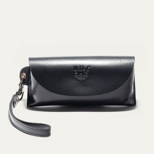 The WILL LEATHER GOODS - EYEGLASS CASE IN BLACK is a handcrafted Italian leather case with a flap closure, embossed logo on the front, and an attached wrist strap on the left, shown against a plain light background.