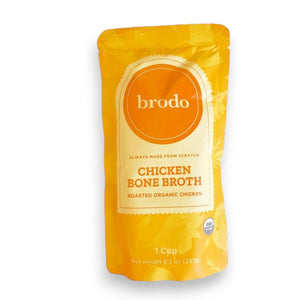 A pouch of BRODO BROTH COMPANY’s BRODO BROTH- CHICKEN BONE BROTH ORGANIC rests on a yellow surface, surrounded by carrots, herbs, and a bowl of collagen-rich broth. The orange packaging with white accents is labeled “Roasted Organic Chicken,” showcasing this keto friendly chicken bone broth as a nourishing addition to any meal.