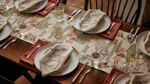 A CHRISTMAS CHEER EMBROIDERED LINEN TABLE RUNNER featuring woodland animals in winter attire and Christmas trees, draped over a wooden dining table set with plates, glassware, and candlesticks.
