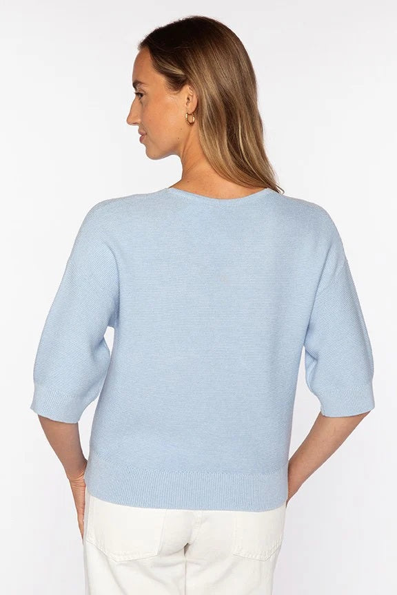 A woman with straight brown hair wears the KINROSS CASHMERE Garter Easy Vee Neck Cotton Sweater in bright pink, featuring a relaxed fit and elbow-length sleeves, paired with white pants against a plain white background.