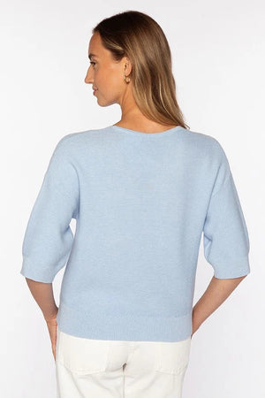 A woman with straight brown hair wears the KINROSS CASHMERE Garter Easy Vee Neck Cotton Sweater in bright pink, featuring a relaxed fit and elbow-length sleeves, paired with white pants against a plain white background.