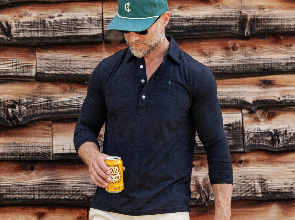 A man with short brown hair, wearing sunglasses, blue jeans, and the CRIQUET BRRR° Long Sleeve Range Polo in Dark Knight by CRIQUET, holds a takeaway coffee cup while standing against a white background.