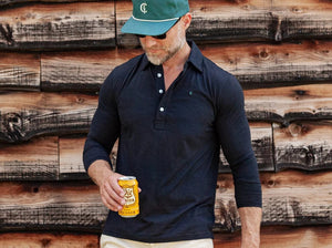 A man with short brown hair, wearing sunglasses, blue jeans, and the CRIQUET BRRR° Long Sleeve Range Polo in Dark Knight by CRIQUET, holds a takeaway coffee cup while standing against a white background.