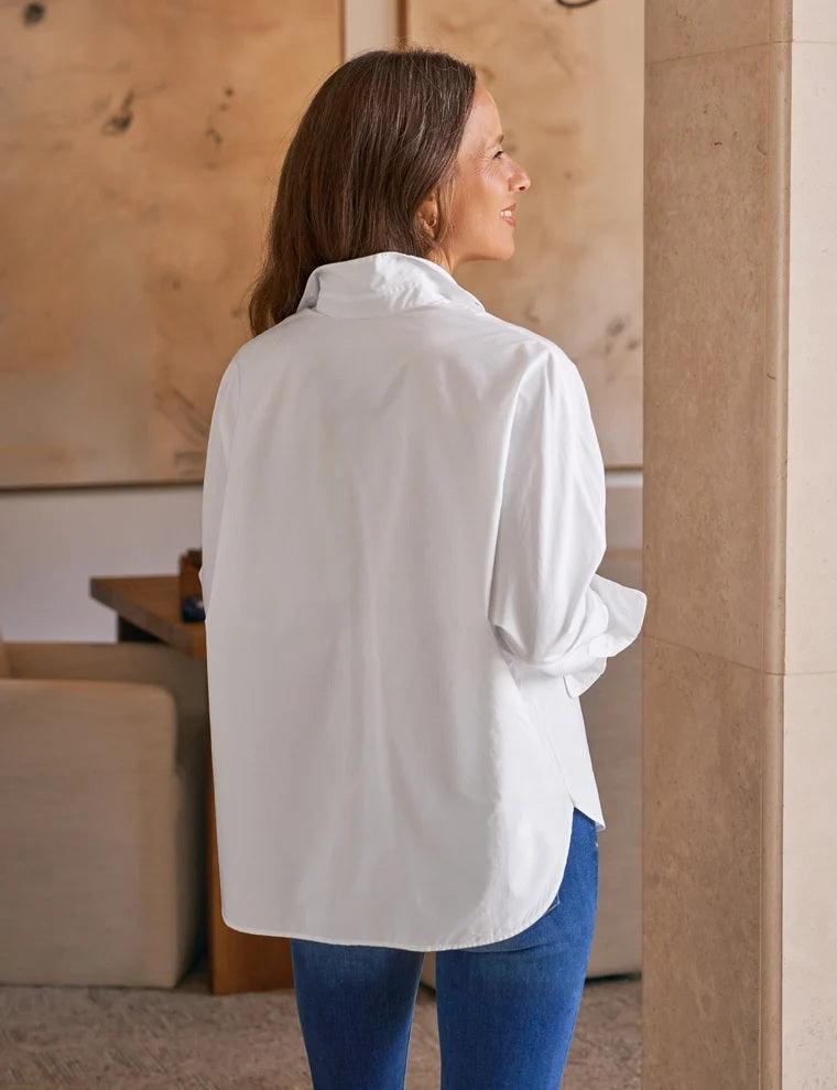A woman smiles while leaning against a beige pillar, wearing the FRANK & EILEEN Oona Continuous Sleeve Button Up Shirt in Regenerative Cotton White and blue jeans, with one hand in her pocket. Neutral-toned artwork and furniture are visible in the background.